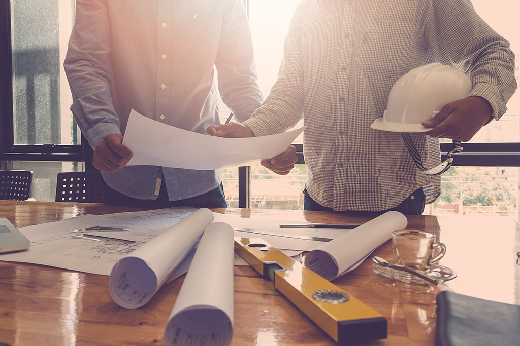an image of two men looking at some blueprints on a tabletop holding their hardhat helmets under their arm